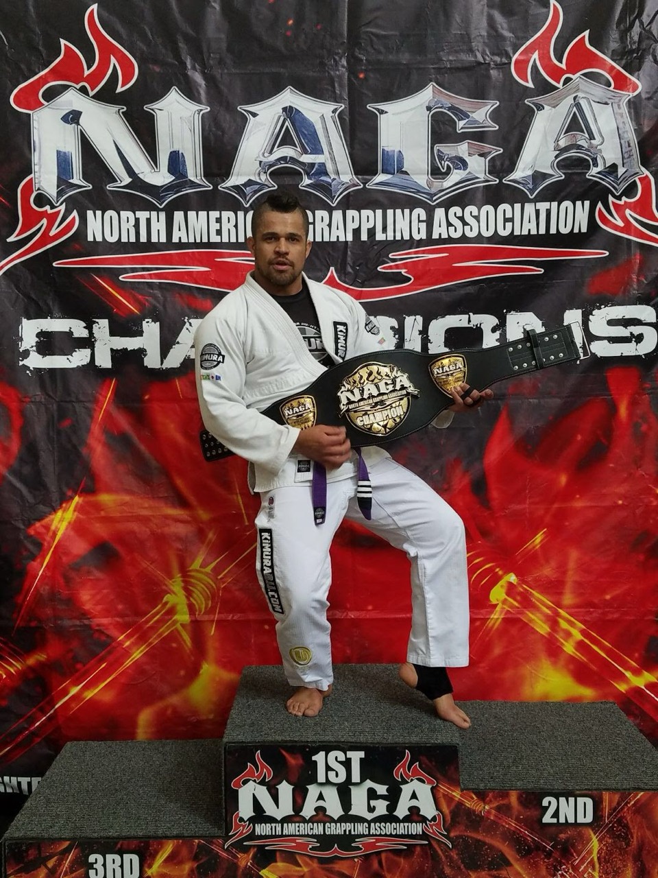 Romulo Caitano Gomes standing on a first-place grappling podium holding a championship belt